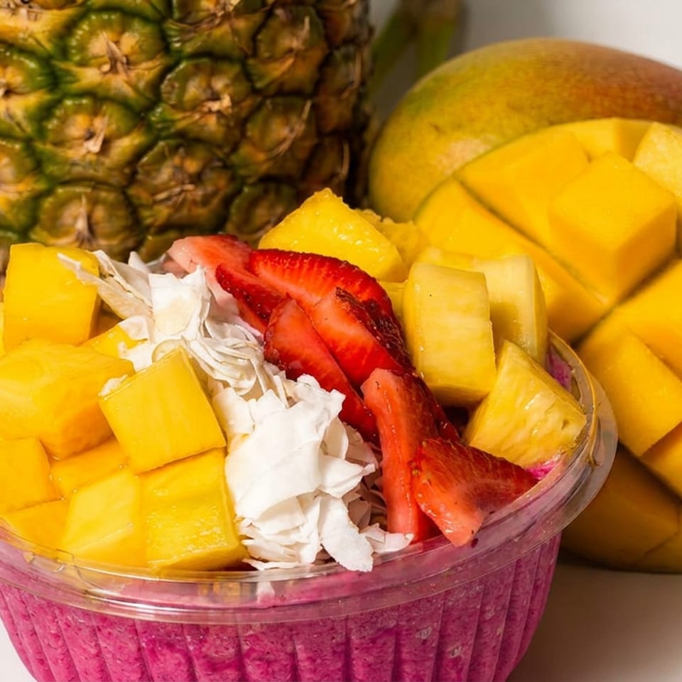 Vibrant fruit bowl topped with mango, pineapple, strawberries, coconut, and cream, with whole pineapple and scored mango behind it.